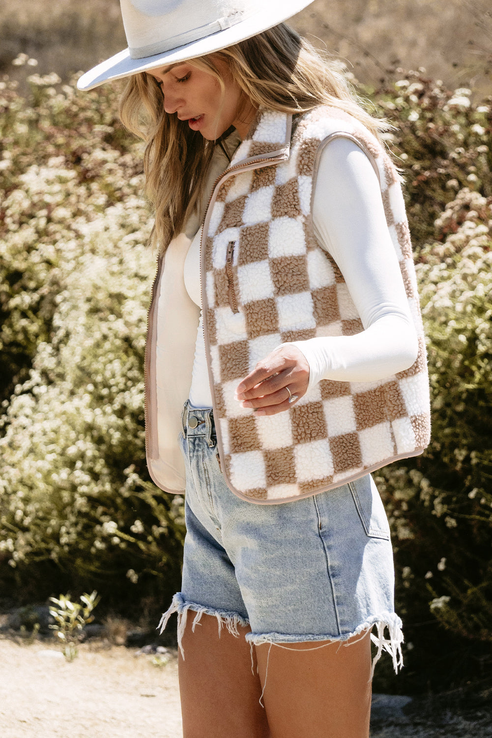 Woman wearing a checkered vest, white shirt, denim shorts, and white hat outdoors.